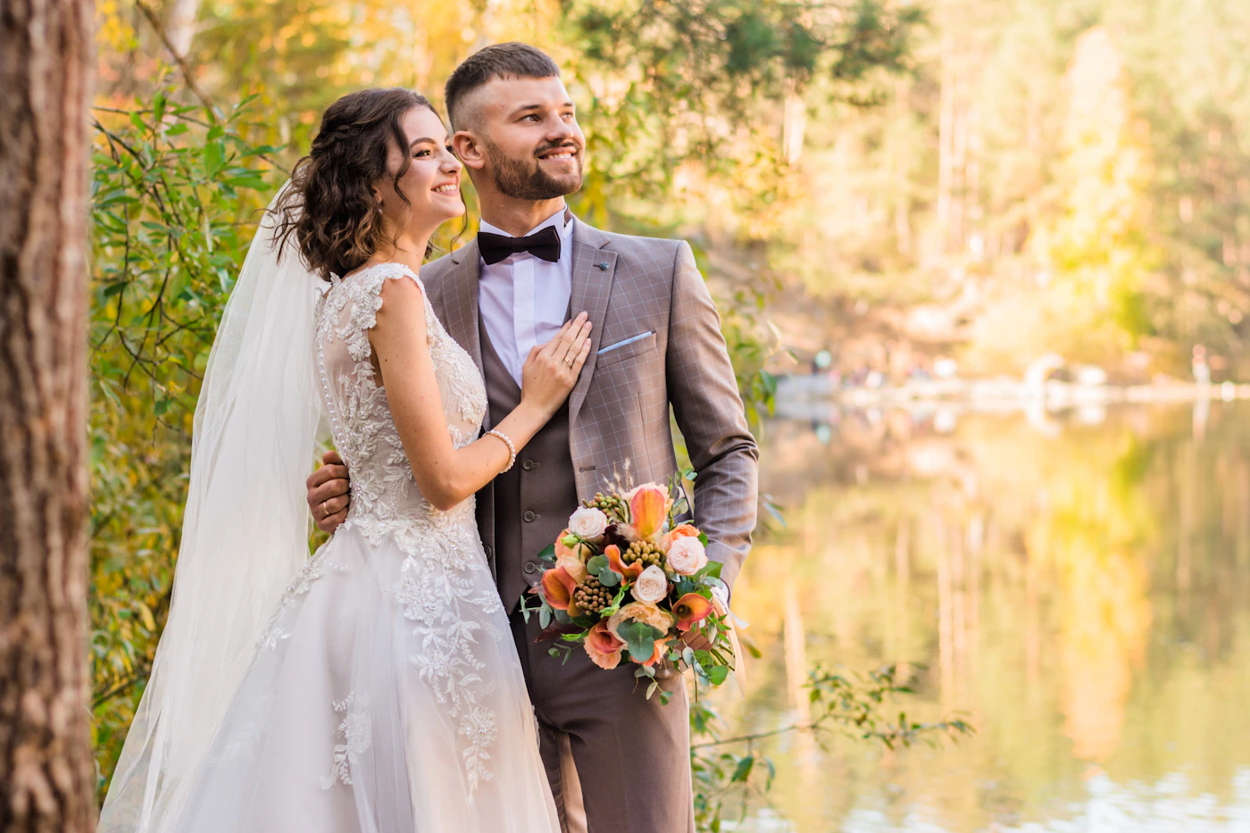 Couple in elegant wedding attire standing in a sunlit forest beside a still lake